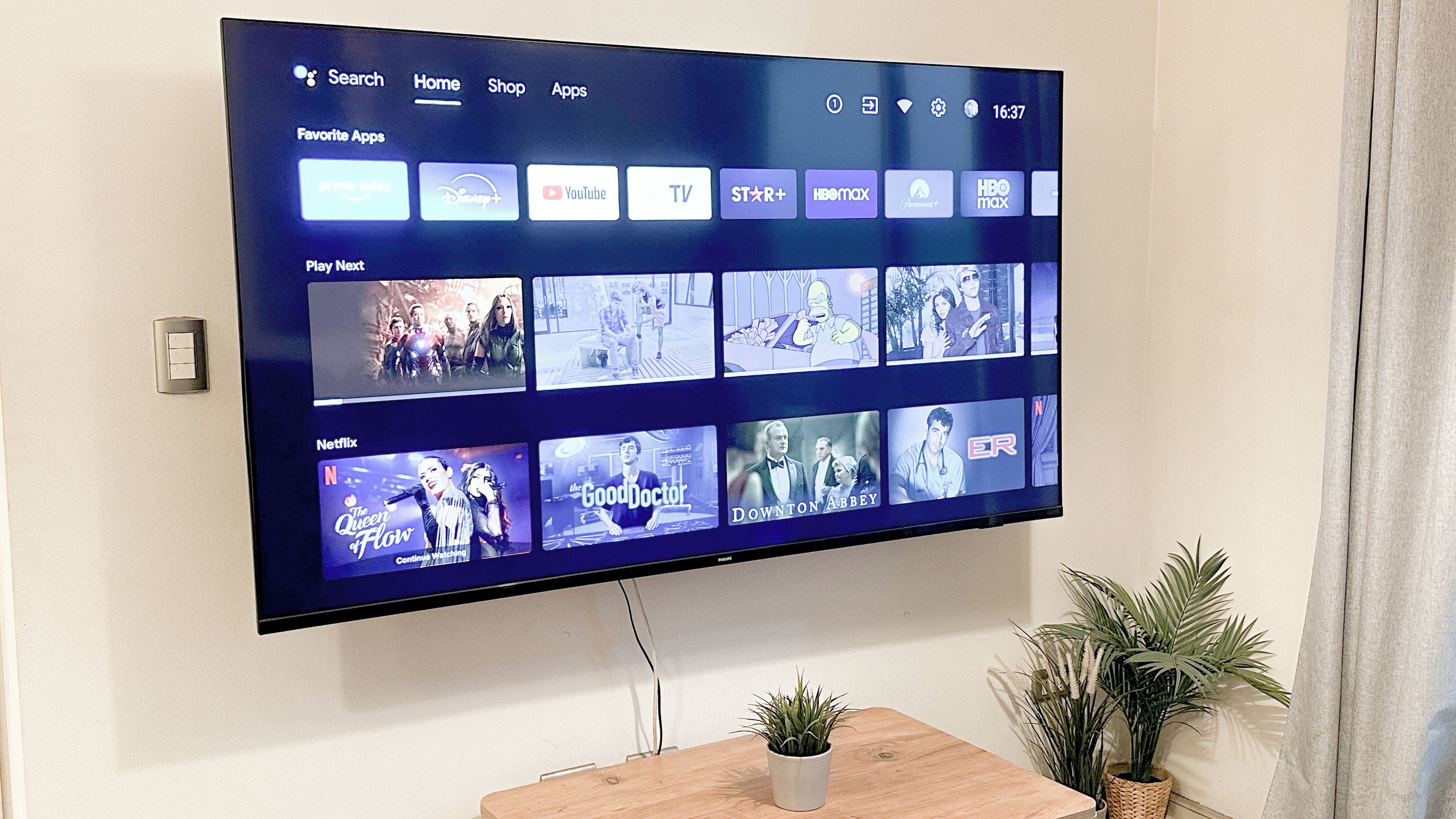 Un televisor de pantalla plana montado en la pared que muestra una interfaz de servicio de transmisión con varios iconos de aplicaciones y una sección 'Reproducir siguiente'.