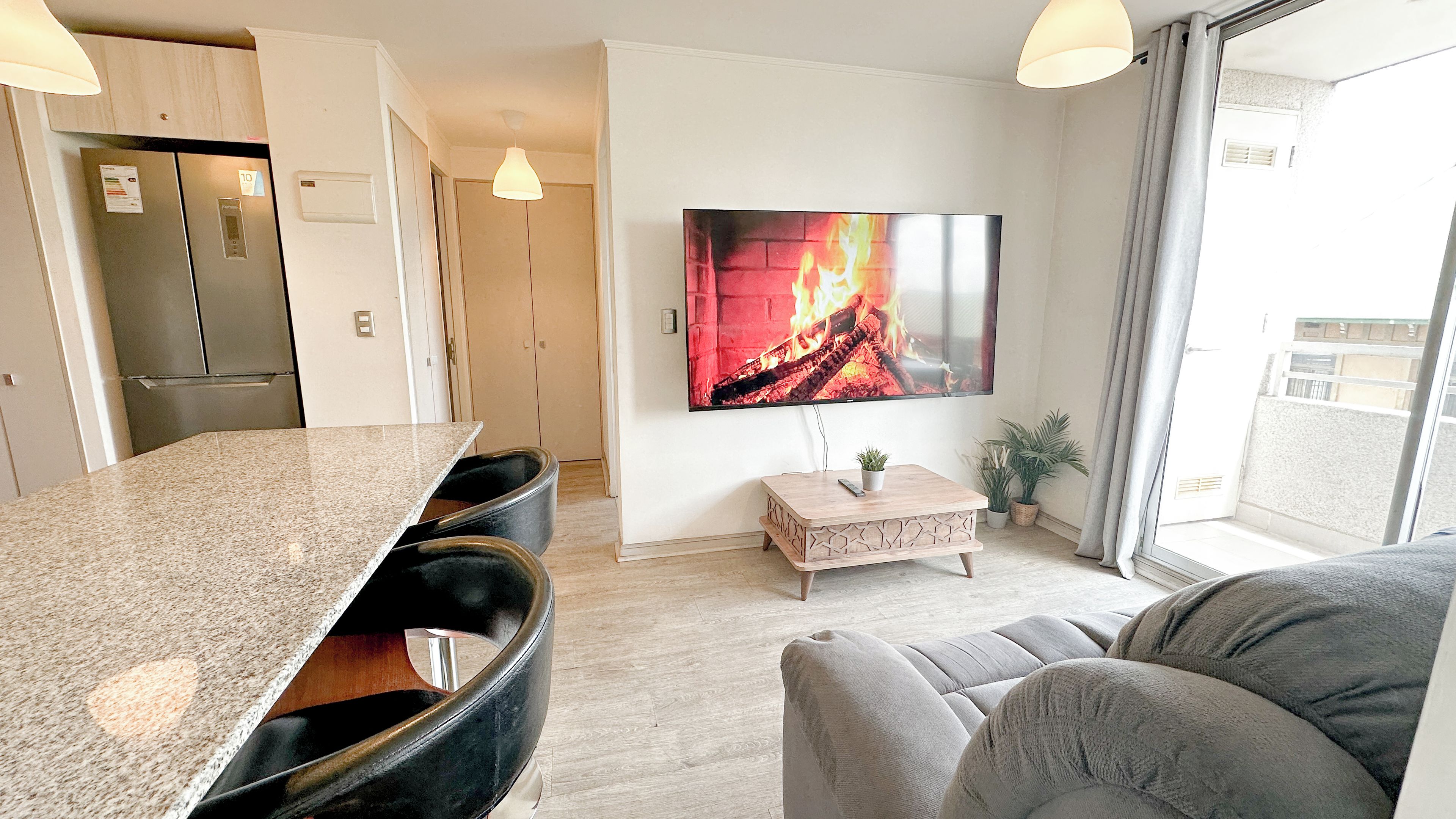 A modern living room with a flat-screen TV, a sofa, a coffee table, and a kitchen area with a refrigerator.