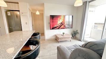A modern living room with a flat-screen TV, a sofa, a coffee table, and a kitchen area with a refrigerator.