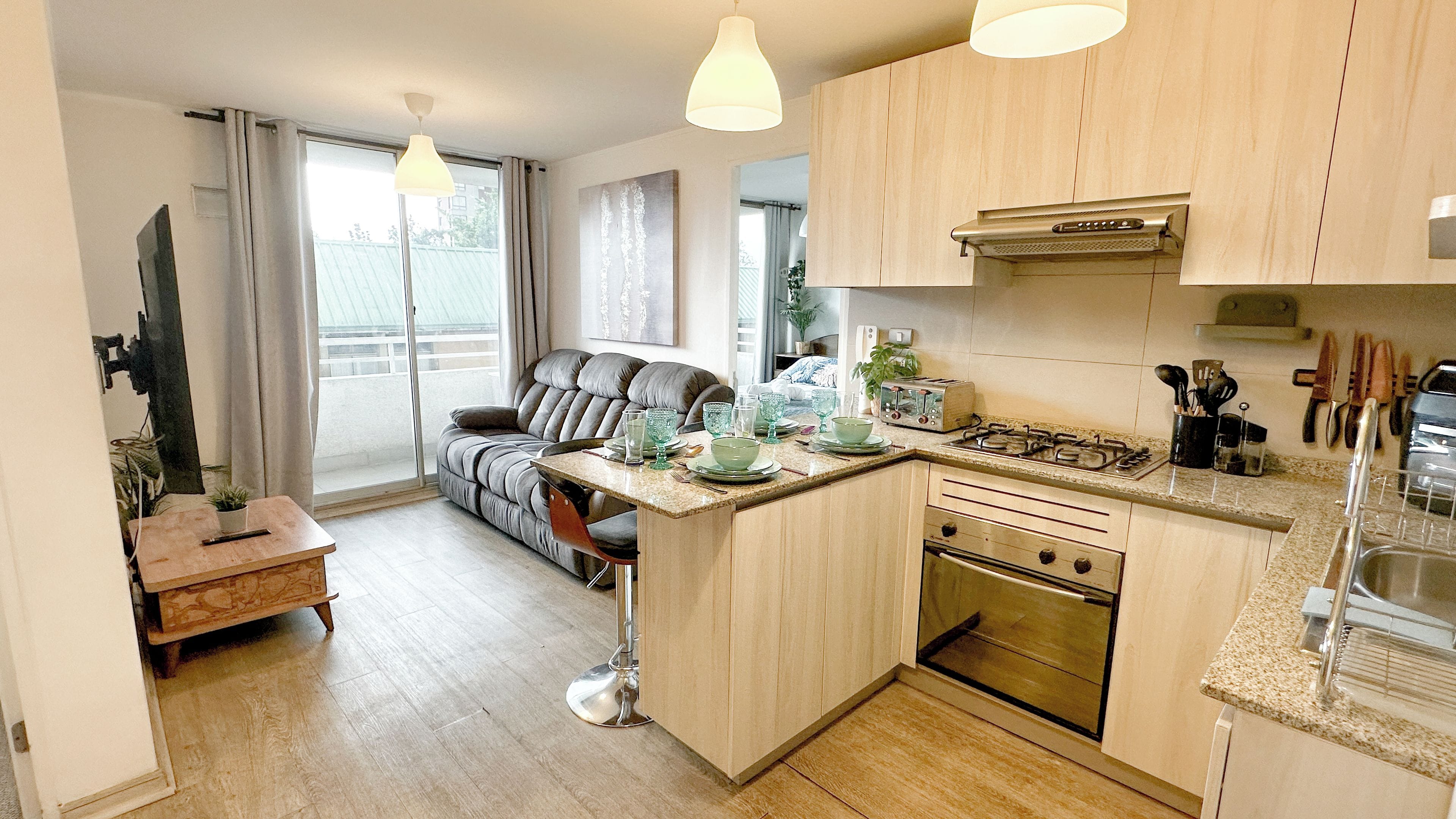 A compact kitchen with a central island, a dining area with a sofa, and a view through a sliding door to a balcony.