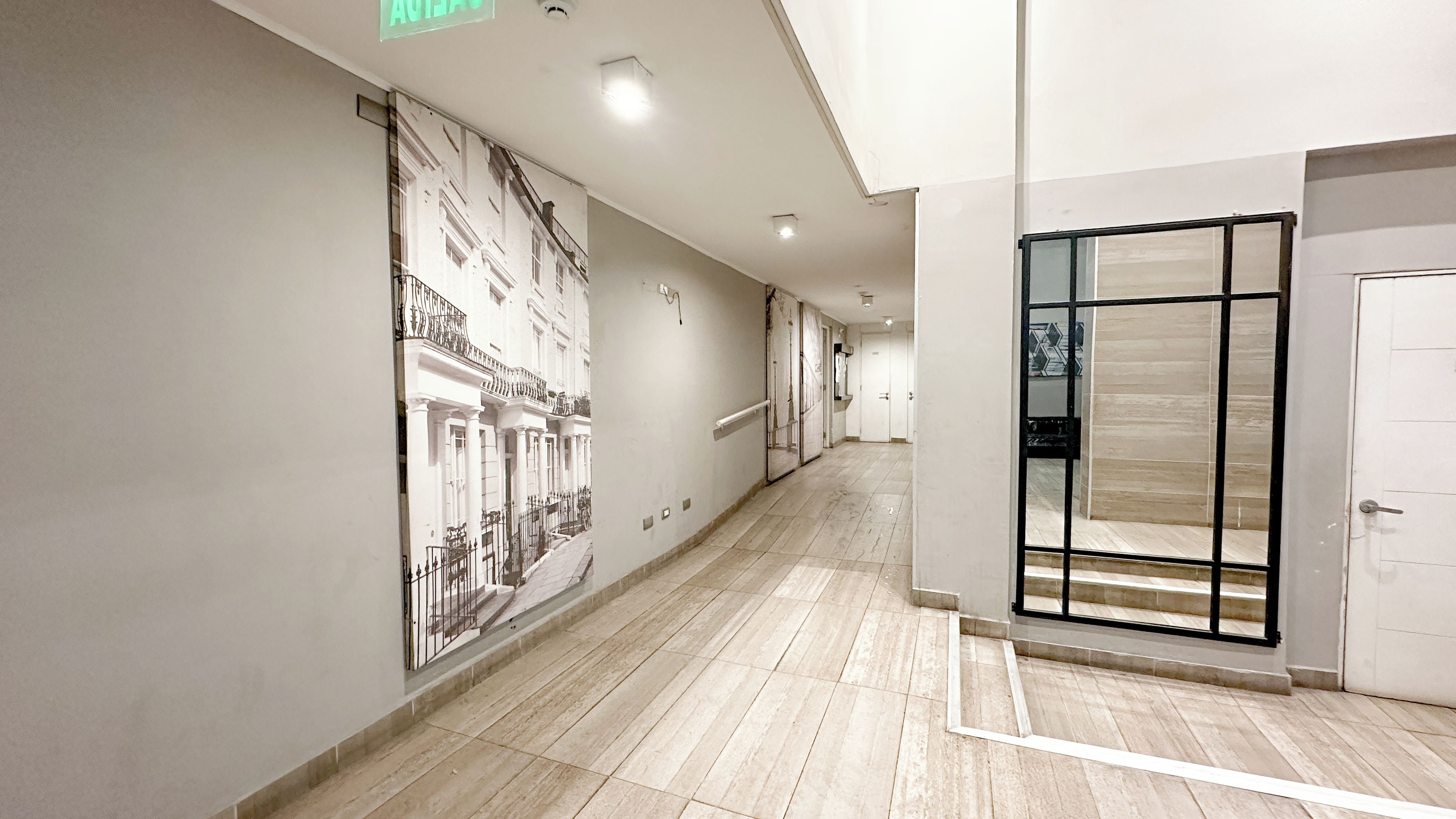 A corridor with a tiled floor, a wall-mounted light fixture, and a door with a black frame.