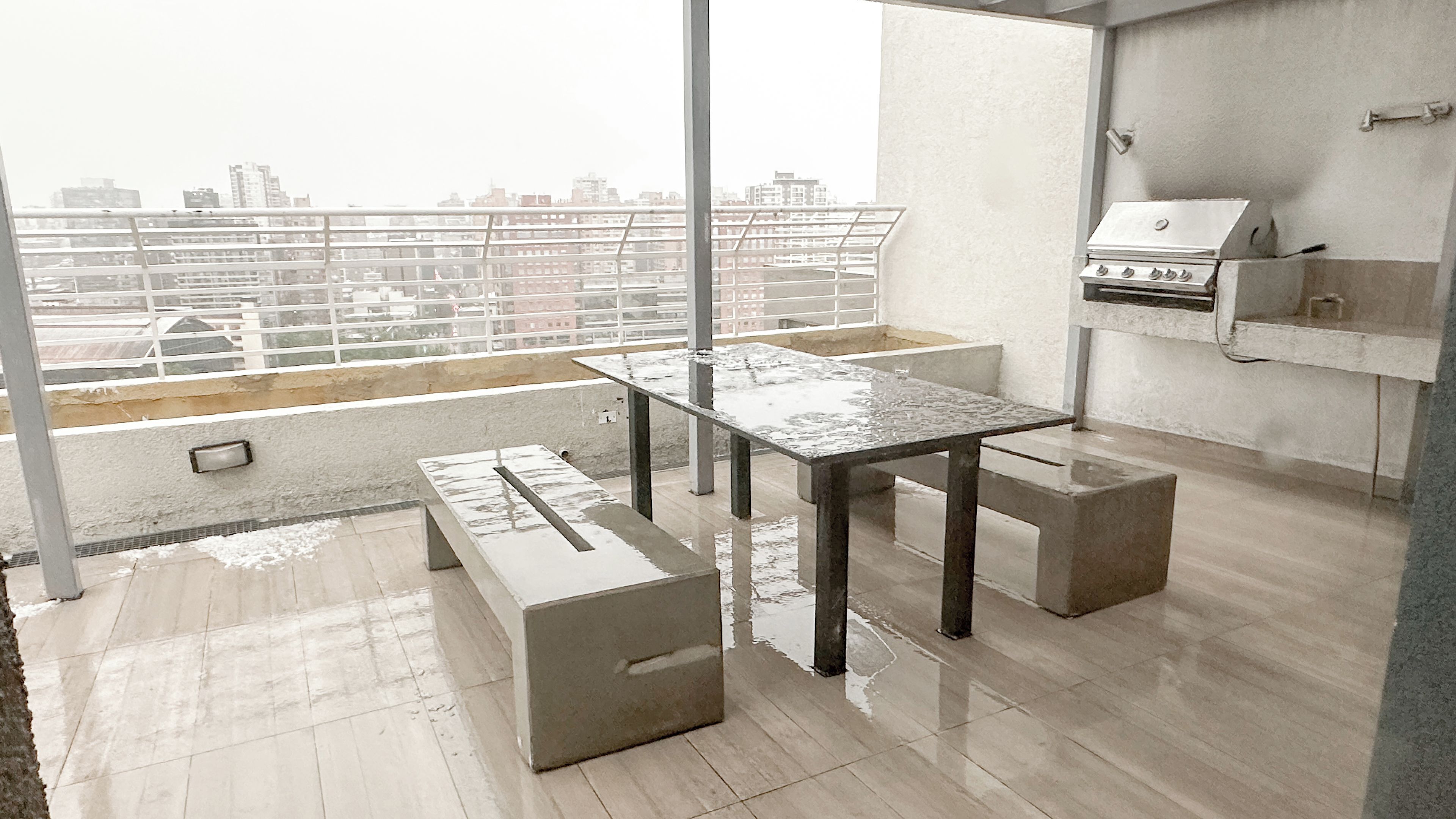 Una terraza en la azotea con mesa de vidrio y bancos, una parrilla y vistas a la ciudad.