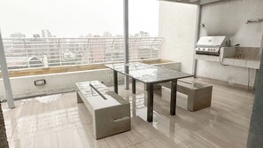 Una terraza en la azotea con mesa de vidrio y bancos, una parrilla y vistas a la ciudad.