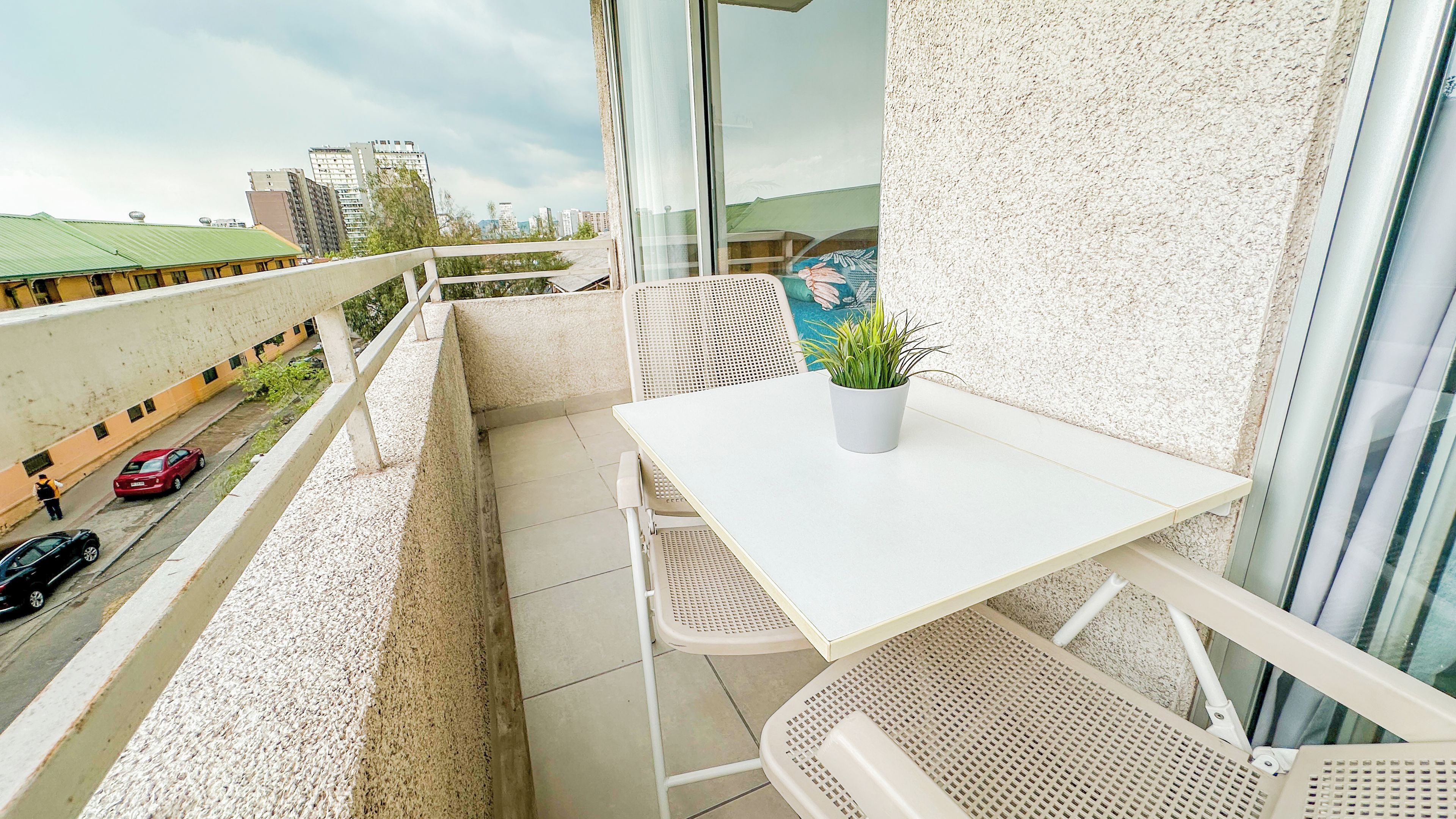Un balcón con una mesa y sillas blancas, una planta en maceta y vista a la calle y edificios.