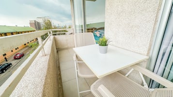 Un balcón con una mesa y sillas blancas, una planta en maceta y vista a la calle y edificios.