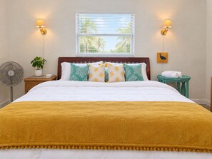 A hotel room with a bed, a window with blinds, two wall-mounted lamps, a bedside table with a plant, a fan, and a small table with towels. - Downtown Venice Island | private patio, walkable (Venice)