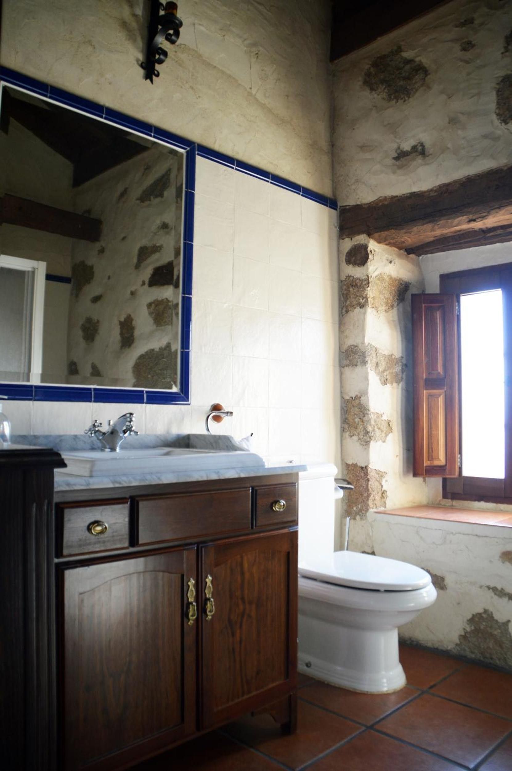 A bathroom with a wooden vanity, a white sink, a mirror, a toilet, and a window with wooden shutters.