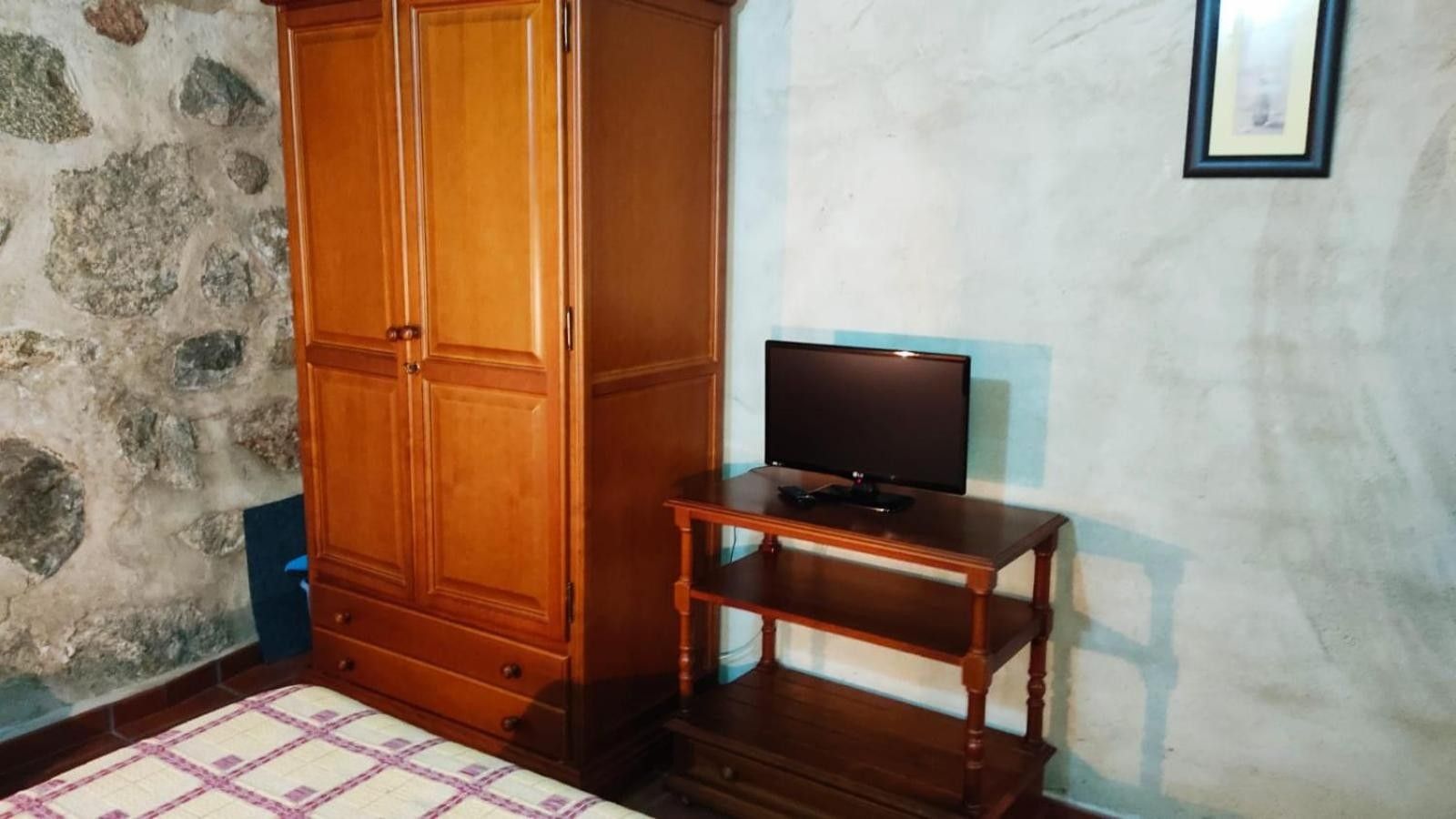 A room with a wooden wardrobe, a television on a wooden stand, and a stone wall.