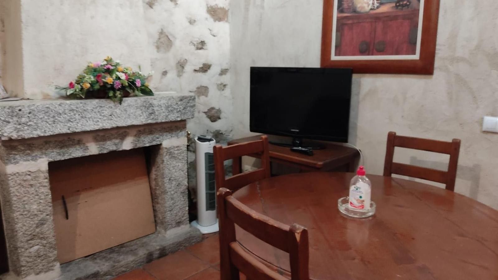 Una habitación con chimenea de piedra, un televisor sobre un mueble de madera, una mesa de comedor con sillas y un cuadro en la pared.