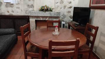 Una mesa de comedor redonda de madera con seis sillas, una lámpara de mesa pequeña y un arreglo floral sobre una repisa de chimenea de piedra.