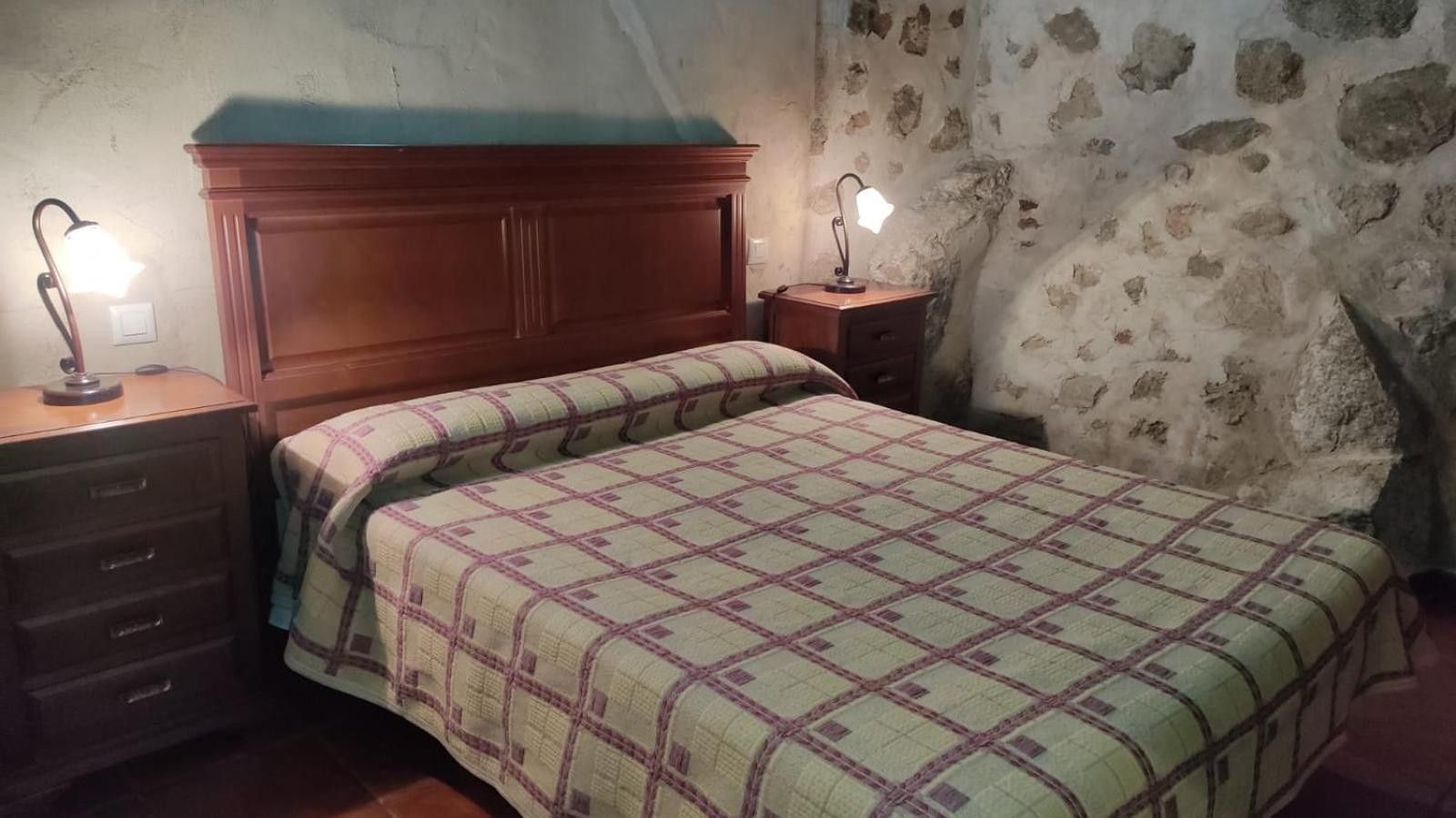 Un dormitorio con cabecera de madera, cama con colcha a cuadros, dos mesitas de noche con lámparas y una pared de piedra.