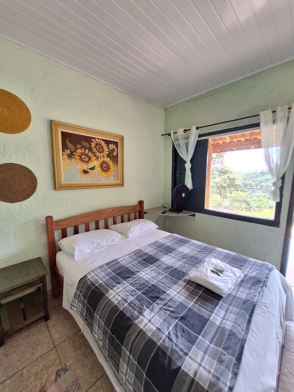A bedroom with a bed, a painting, a window with curtains, and a small table.