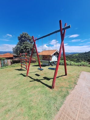 A playground with swings and a slide, surrounded by a grassy area and a building with a tiled roof.