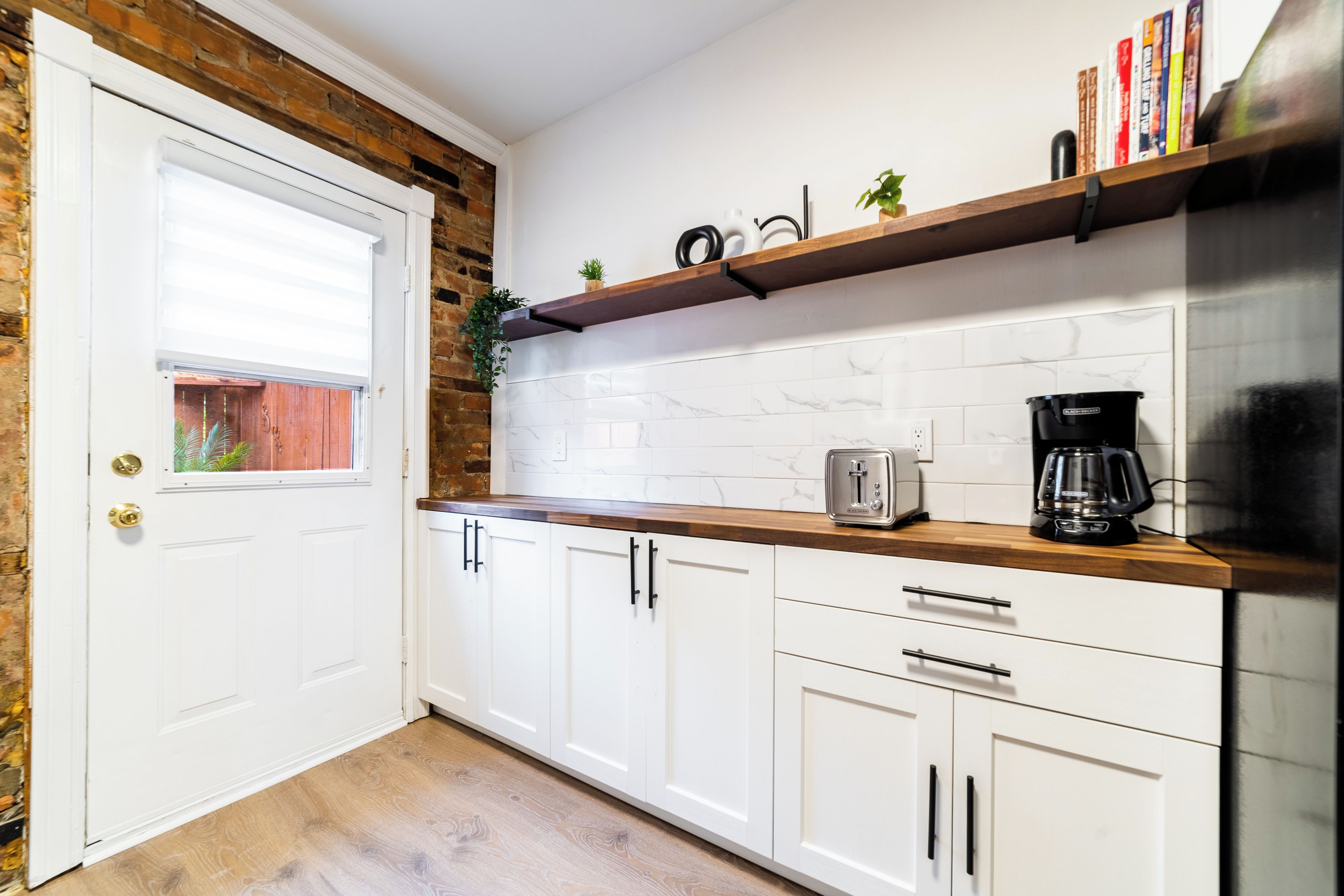 A kitchen with white cabinets, a wooden countertop, a toaster, a coffee maker, and a shelf with books and a plant.