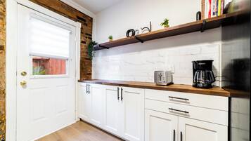 A kitchen with white cabinets, a wooden countertop, a toaster, a coffee maker, and a shelf with books and a plant.
