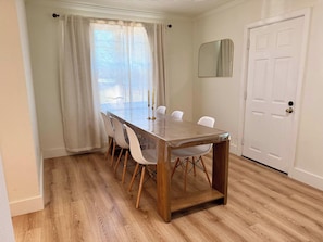 Een eetgedeelte met een houten tafel en witte stoelen, een raam met dunne gordijnen, en een witte deur met een messing deurklink.