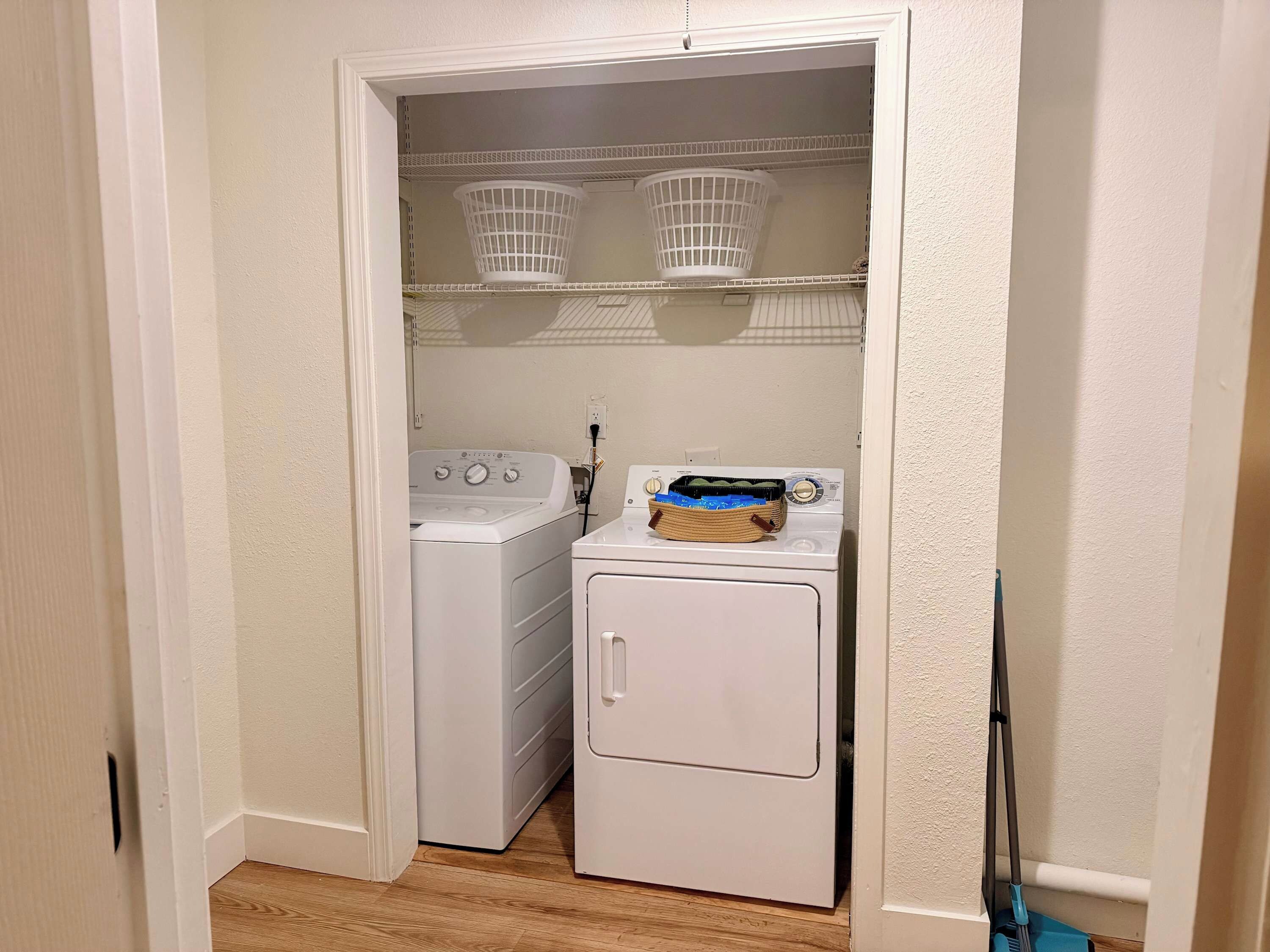 Een wasserij met een wasmachine en droger, twee witte manden op planken, en een dweil die tegen de muur staat.