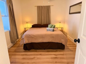 Een slaapkamer met een bed, nachtlampjes, een raam met gordijnen, een nachtkastje en een deur.