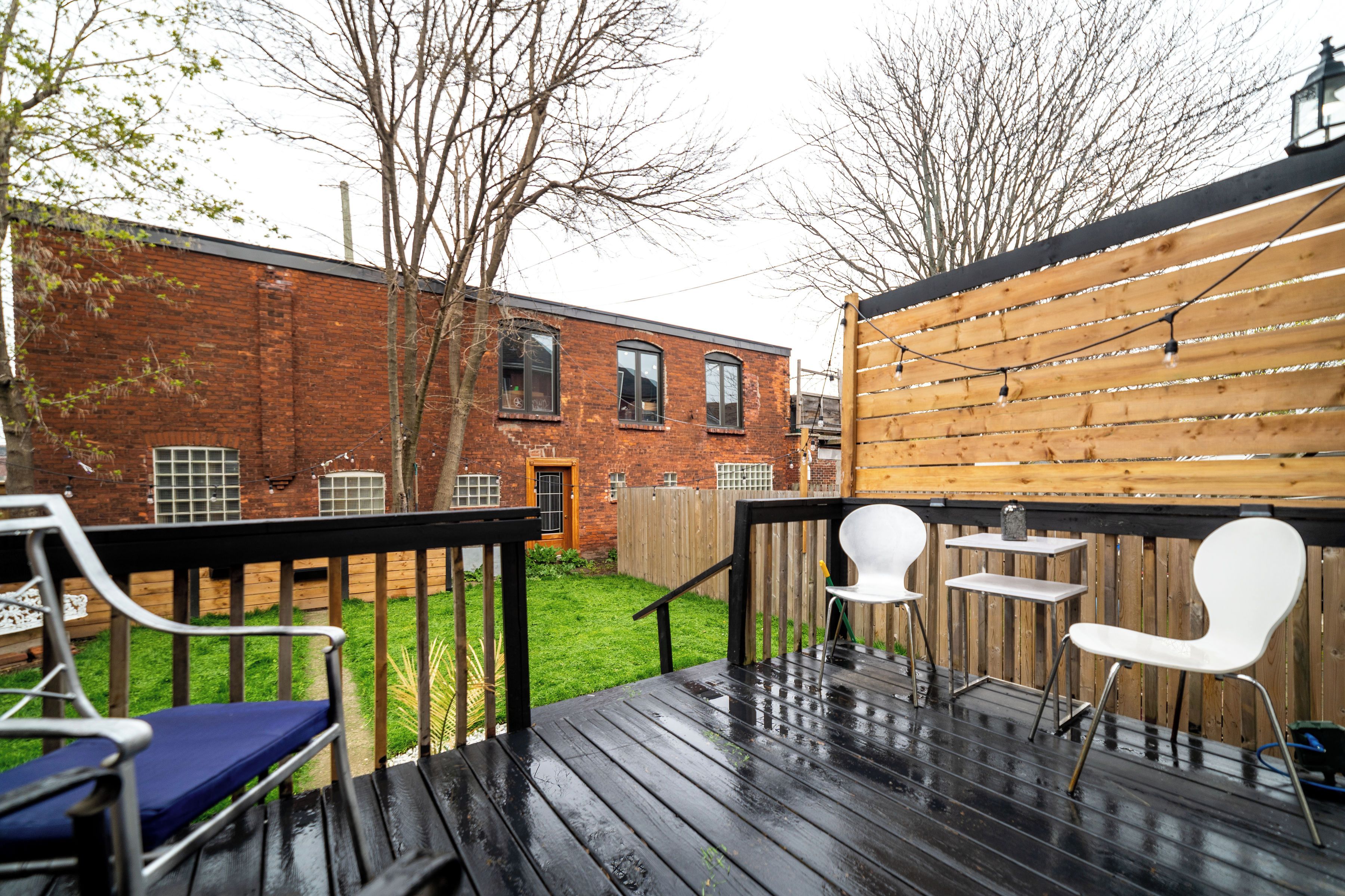 A wooden deck with two white chairs and a table, a brick building in the background, and a wooden fence.