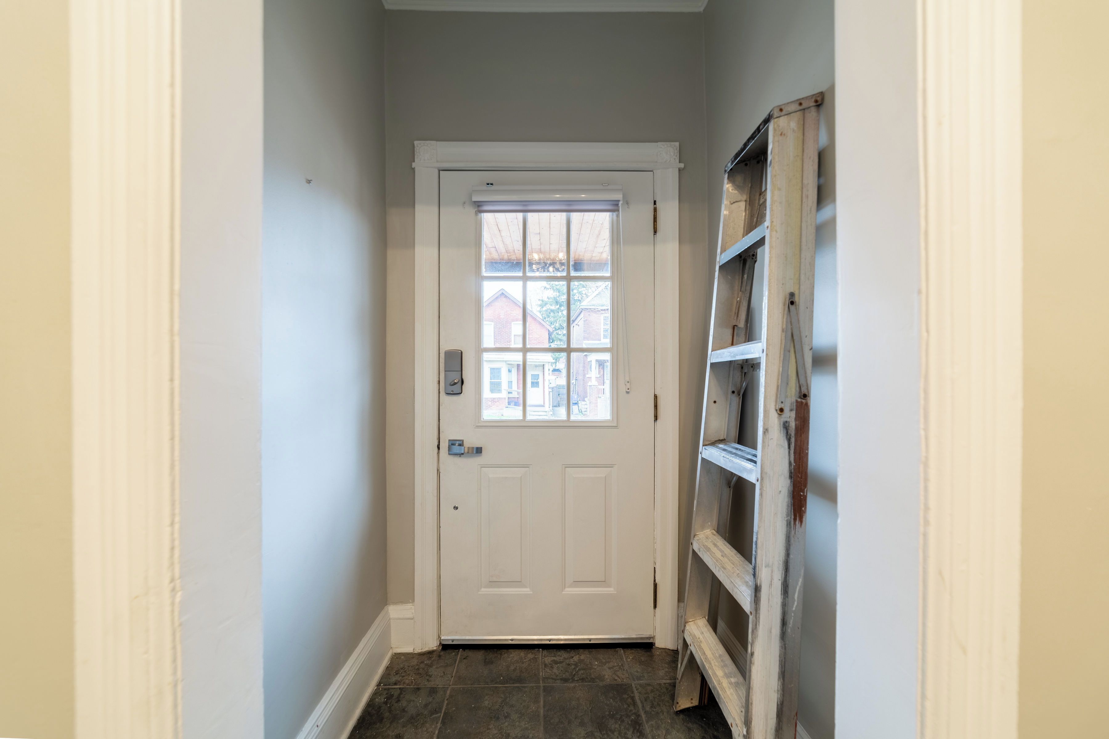 Un couloir étroit avec une porte blanche, une petite fenêtre et une échelle en bois appuyée contre le mur.