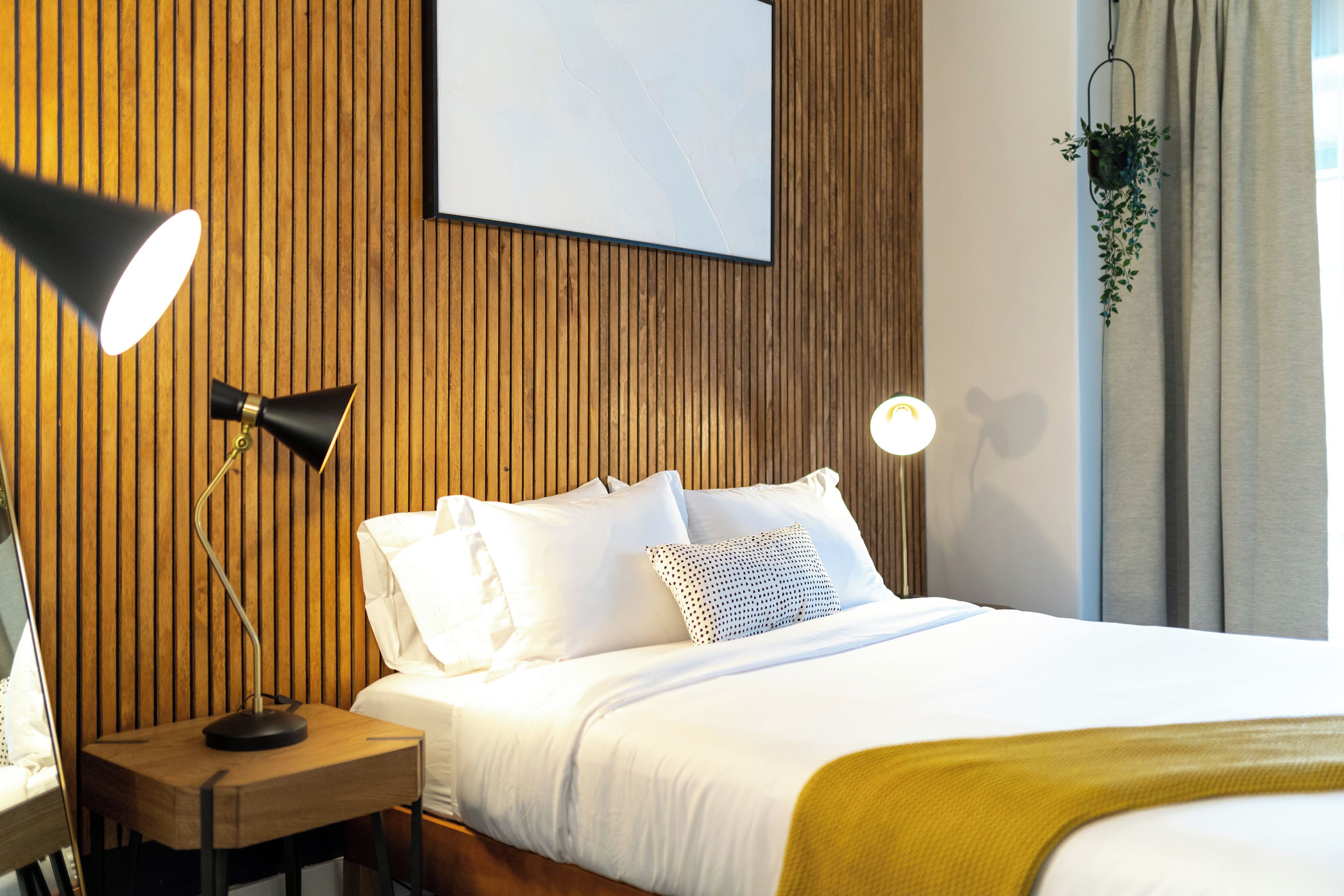Un lit bien fait, avec du linge de lit blanc et un plaid couleur moutarde, une lampe de chevet et un luminaire fixé au mur.