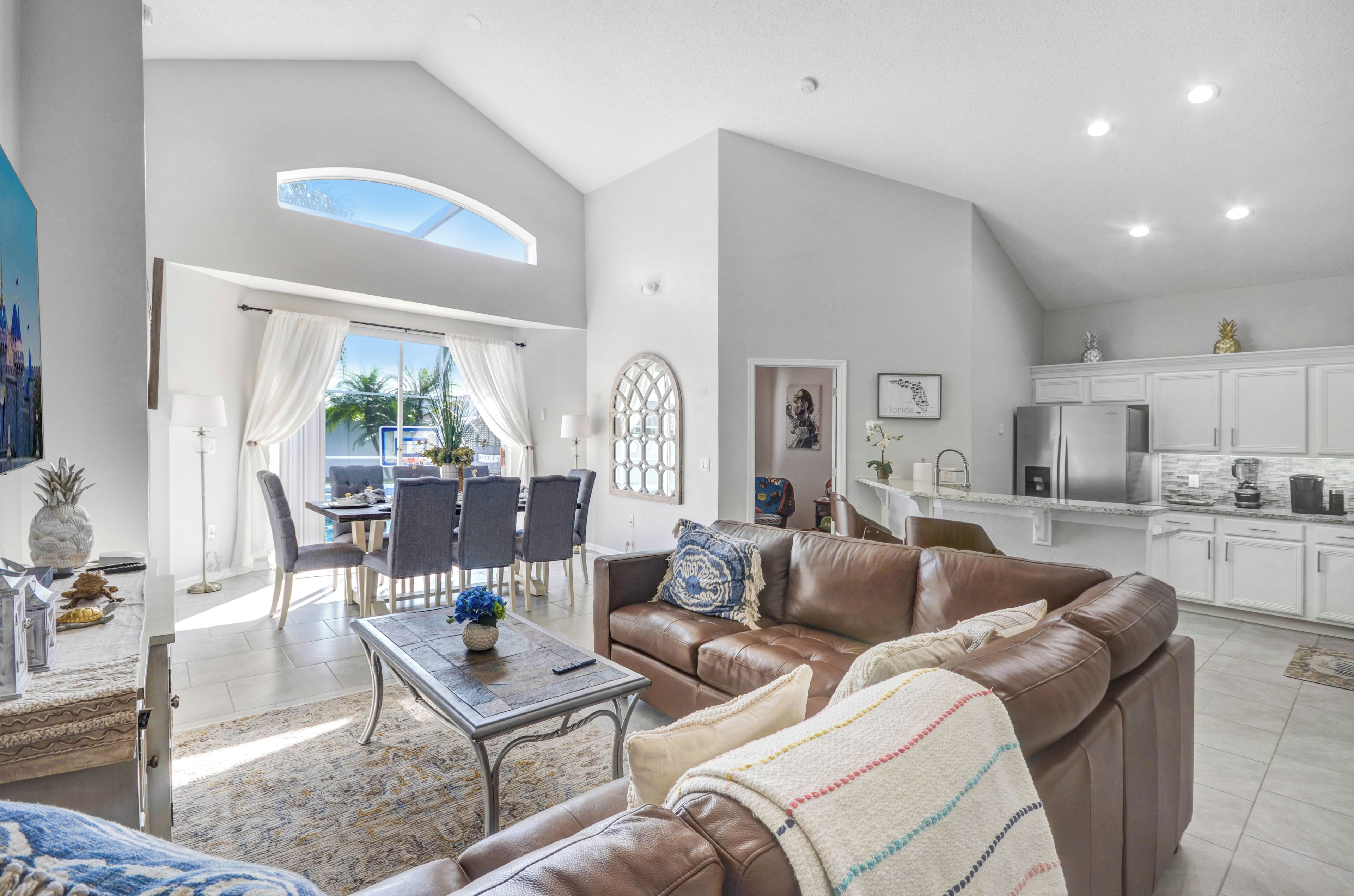 A spacious living area with a brown leather sofa, a dining table with chairs, and a kitchen with white cabinets.