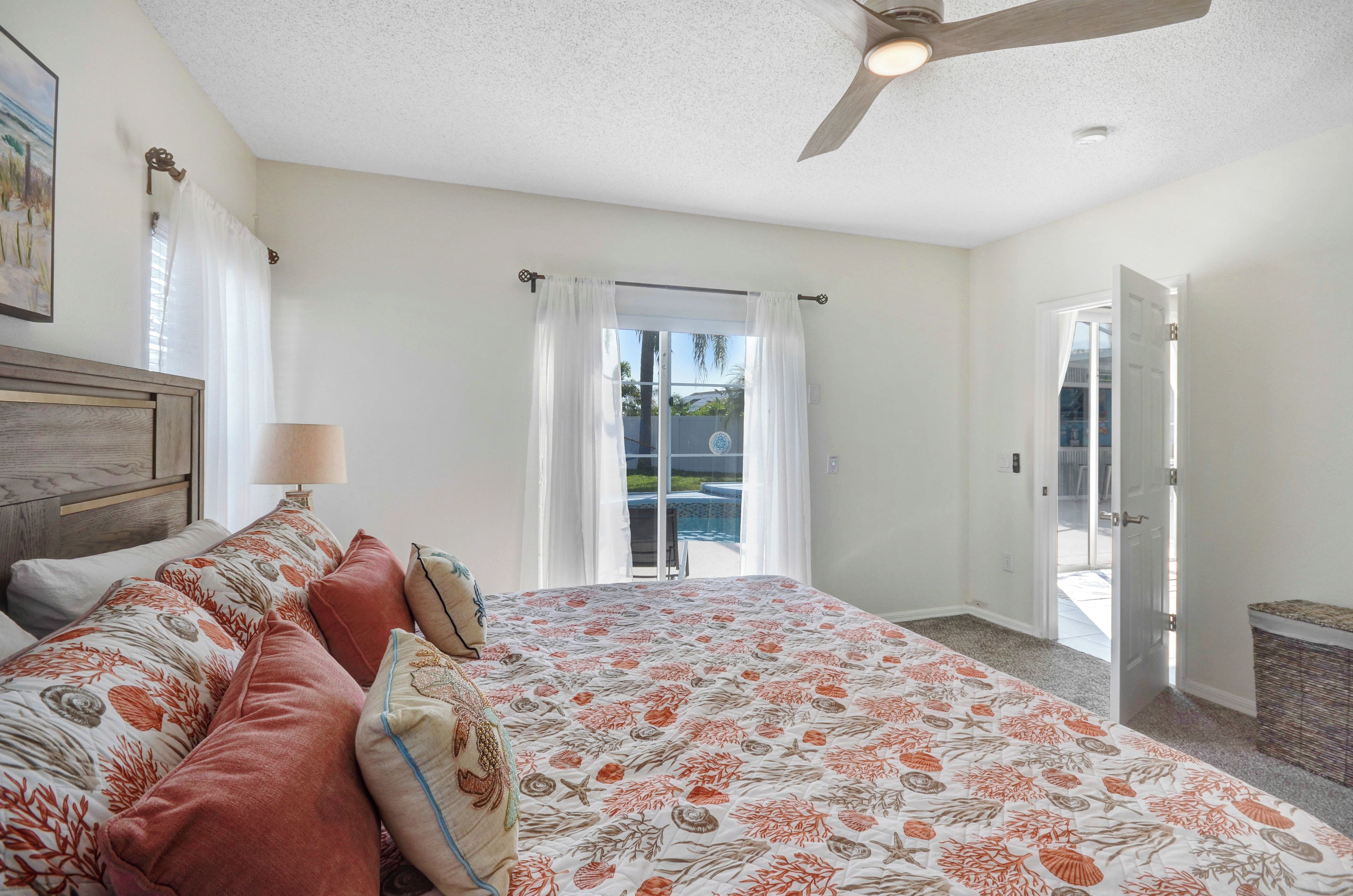 Une chambre avec un lit, une table de chevet, une lampe, un ventilateur de plafond et une vue sur une piscine, visible à travers une porte vitrée.