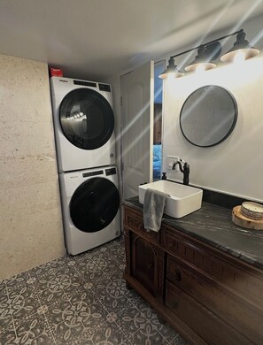 A laundry room with a front-loading washer and dryer, a sink with a mirror above, and patterned floor tiles.