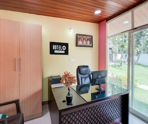 A hotel reception desk with a framed picture, a sign indicating it's a company-serviced hotel, a printer, and a potted plant. - Classic Kaveri Homestay (Shrirangapattana)