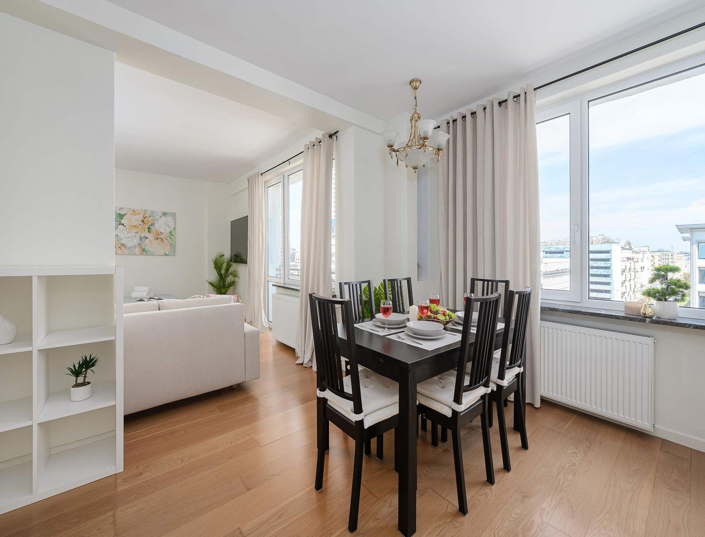 A dining area with a table set for four, surrounded by chairs, a chandelier, and large windows with curtains.