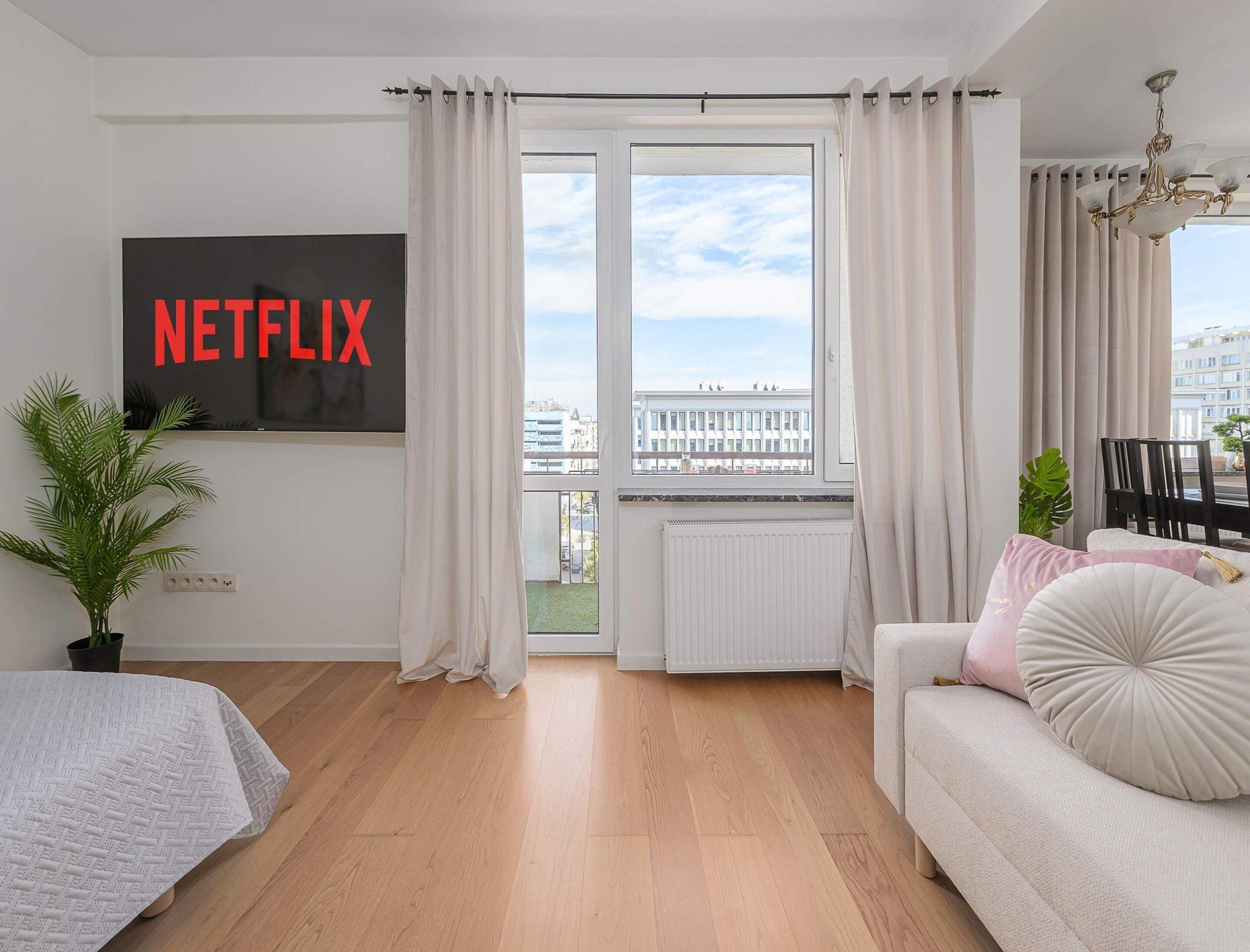 A modern living room with a large window, a sofa, a TV mounted on the wall, and a potted plant.