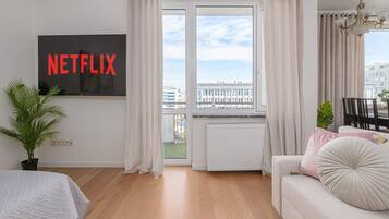 A modern living room with a large window, a sofa, a TV mounted on the wall, and a potted plant.