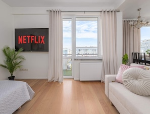 A modern living room with a large window, a sofa, a TV mounted on the wall, and a potted plant. - Apartment With Skyline Balcony (Warsaw)