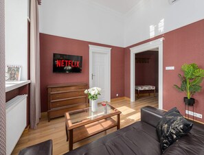 Un salon avec une table basse en bois, un canapé en cuir noir, une porte blanche et un téléviseur fixé au mur qui affiche le logo « NETFLIX ».