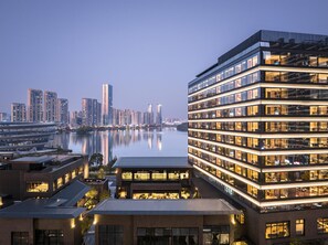 Un hotel moderno con varios pisos, amplias ventanas y vistas a un horizonte urbano sobre un cuerpo de agua.