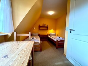 Een kleine, knusse kamer met een schuine kap, een houten tafel, twee eenpersoonsbedden, een plank met een lamp en een deur.