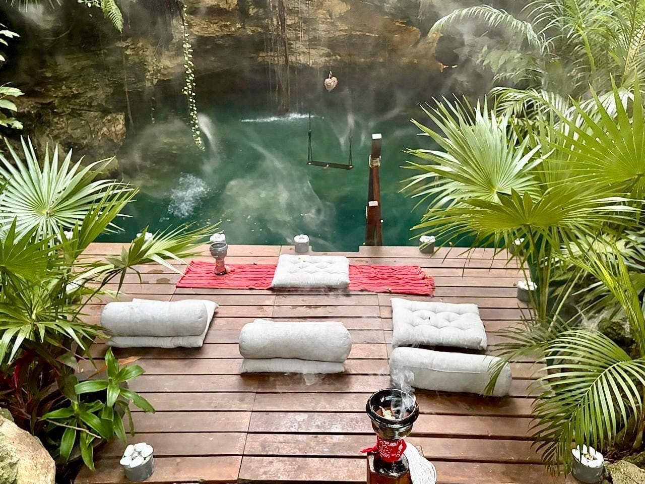 A wooden deck with white cushioned seating and a smoking hookah, overlooking a misty water feature.