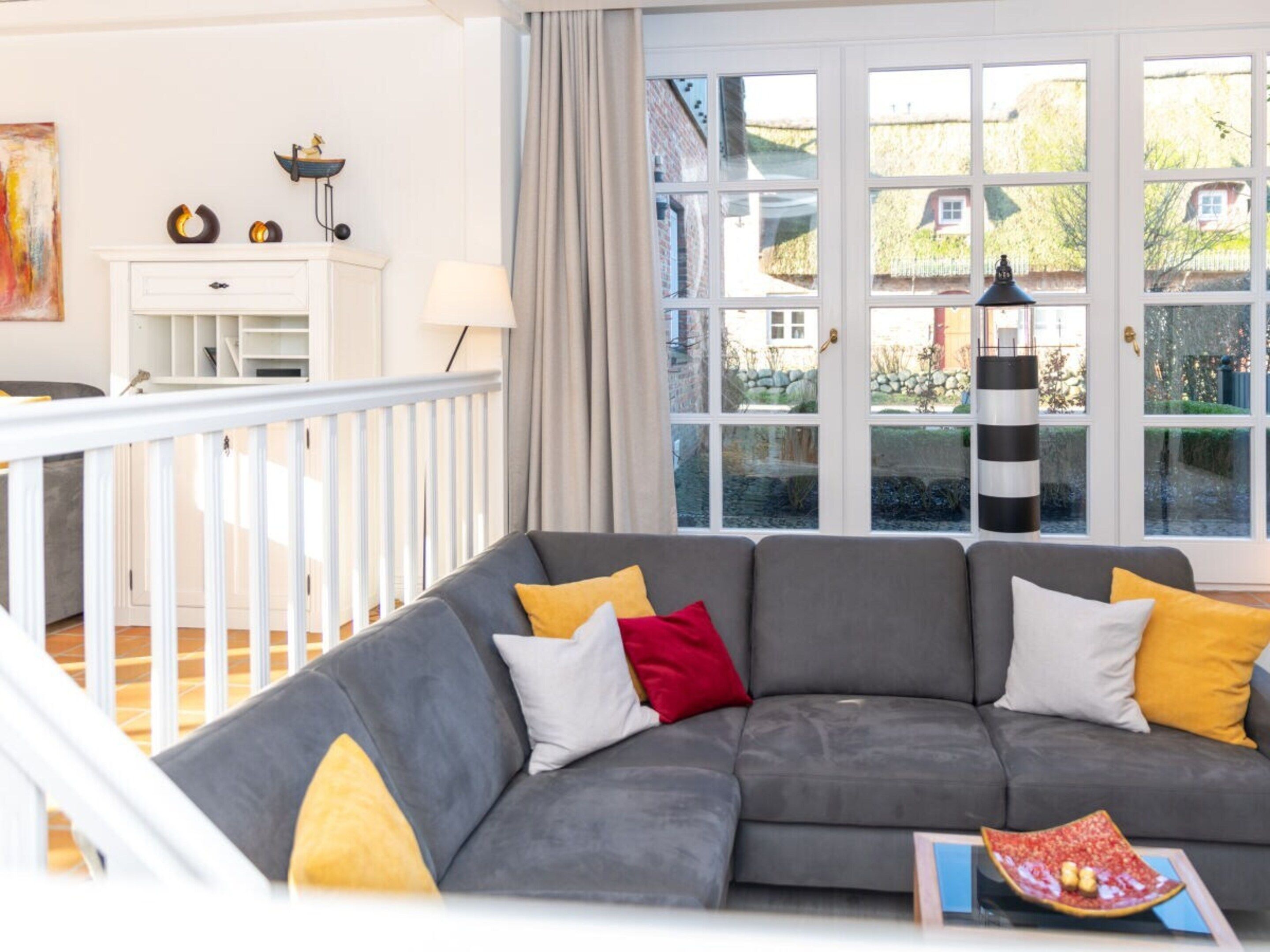 Uma sala de estar com sofá cinzento, almofadas amarelas e vermelhas, aparador branco e uma grande janela com vista para uma casa e jardim.