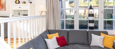 Uma sala de estar com sofá cinzento, almofadas amarelas e vermelhas, aparador branco e uma grande janela com vista para uma casa e jardim.