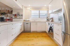 A modern kitchen with white cabinetry, a stainless steel refrigerator, and a dishwasher. - Crafted with Soul A Leafy Boho Escape by the Sea (Port Melbourne)