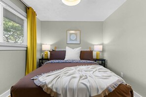 Ein Hotelzimmer mit einem Bett, zwei Nachttischen, einem Fenster mit gelben Vorhängen und einem Wandbild.