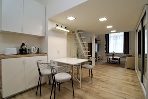 A modern kitchen with white cabinetry, a dining area with a white table and metal chairs, a wooden floor, and a staircase leading to a living area with a sofa and a TV. - Roxy Stúdió 4A - Budapest (Budapest)