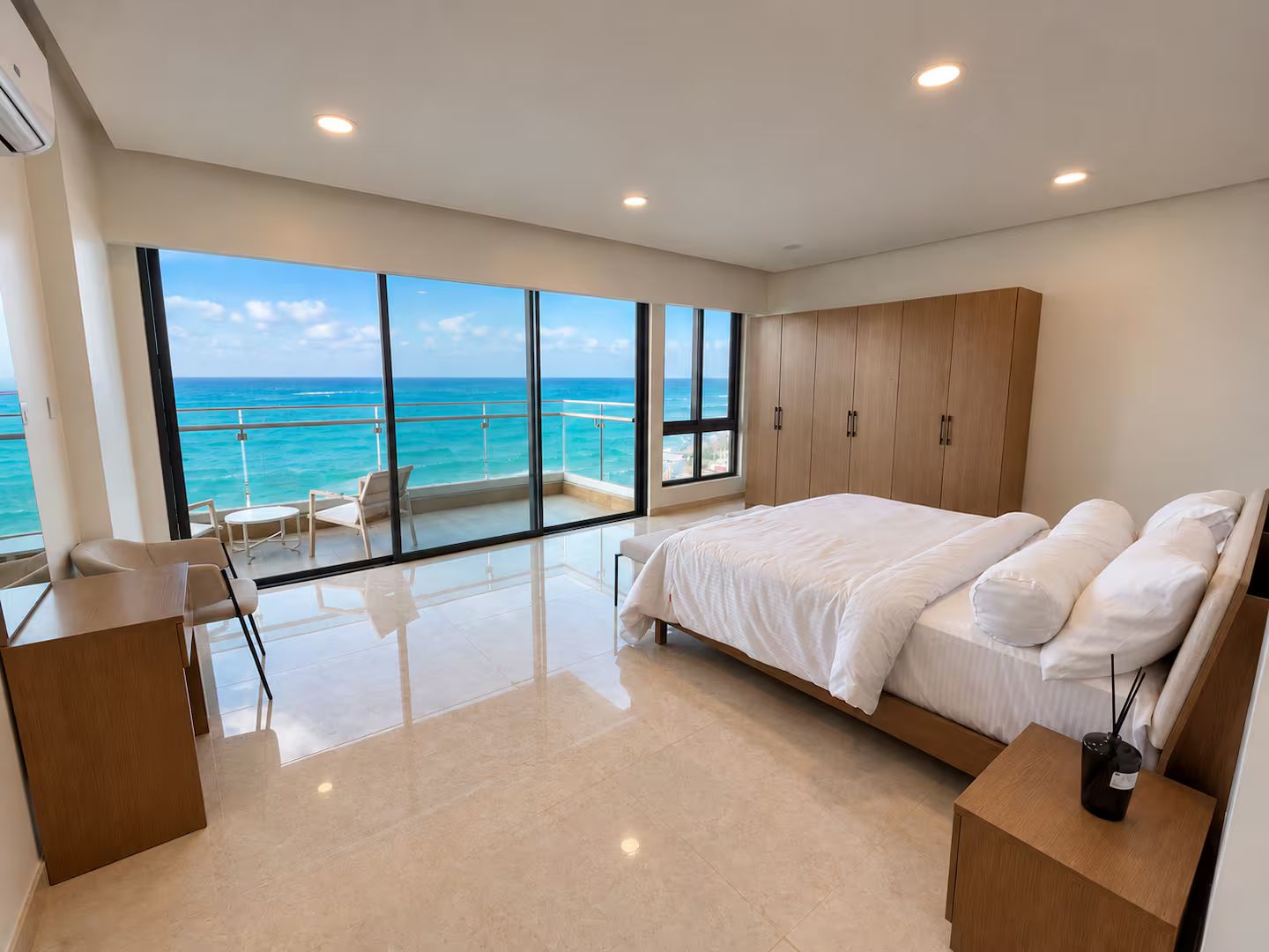 A modern bedroom with a large bed, a balcony with a view of the ocean, and a seating area with a table.
