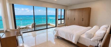 A modern bedroom with a large bed, a balcony with a view of the ocean, and a seating area with a table.