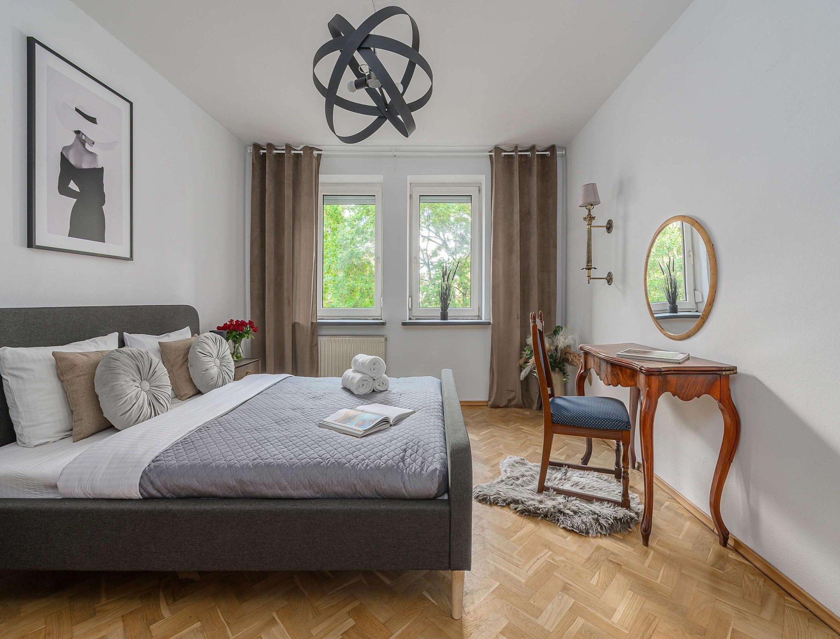 A bedroom with a bed, a chair, a desk, a mirror, and two windows with curtains.