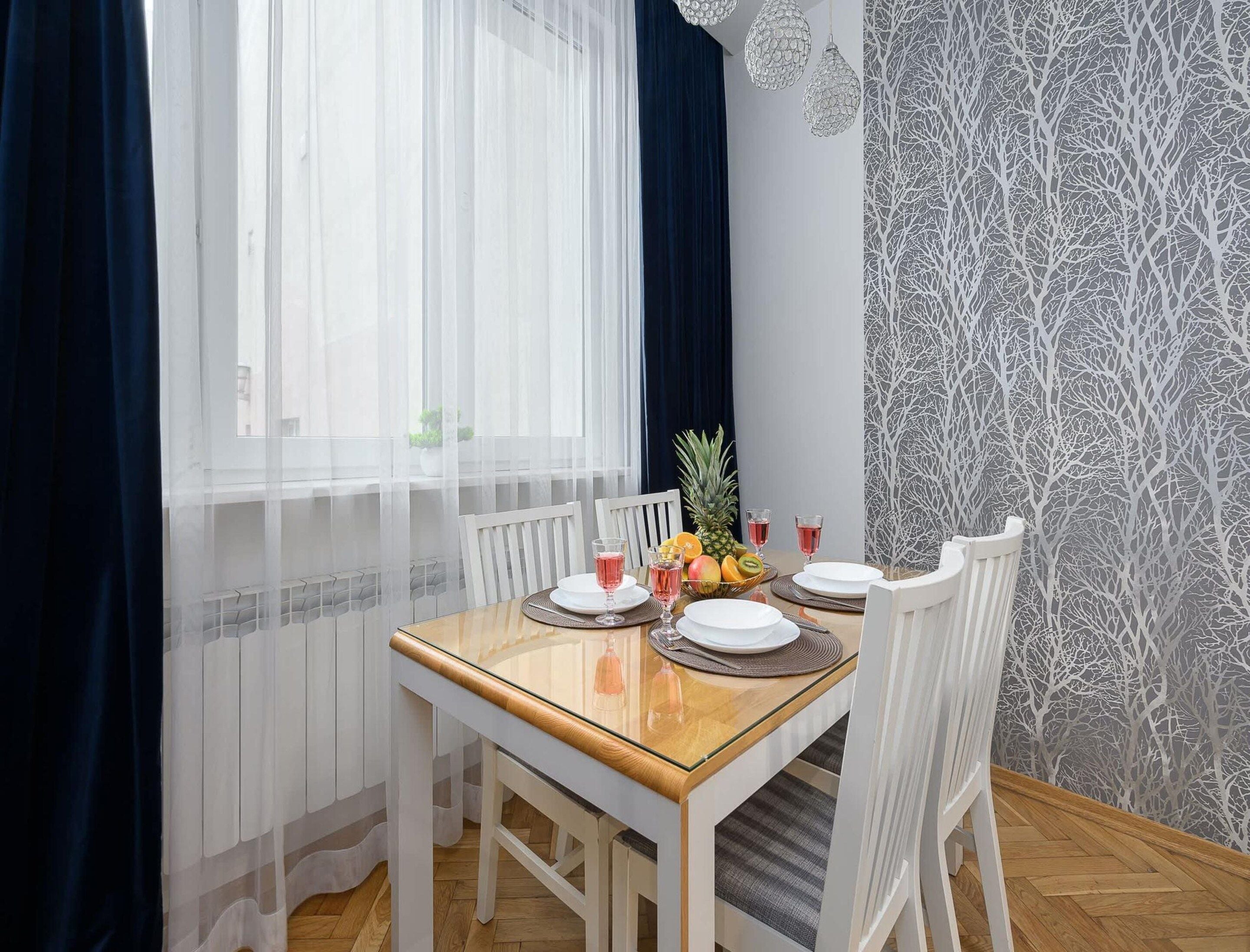 A dining area with a table set for two, white chairs, a fruit bowl, and a window with sheer curtains.