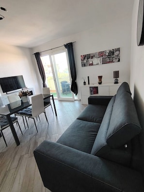 A modern living room with a sofa, a dining table with chairs, a TV, and a large window with curtains. - New Listing (Porto Torres)