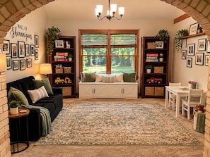 A cozy living room with a black sofa, a white bench, and a patterned rug. There are bookshelves filled with books and toys, and a wall with framed pictures. - Wooded Acre Retreat near Wright-Patt w/ Game Room (Xenia)
