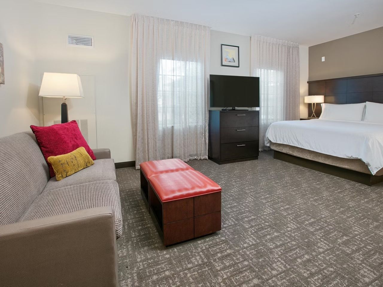 A hotel room with a bed, a sofa, a TV, a desk, and a red ottoman.