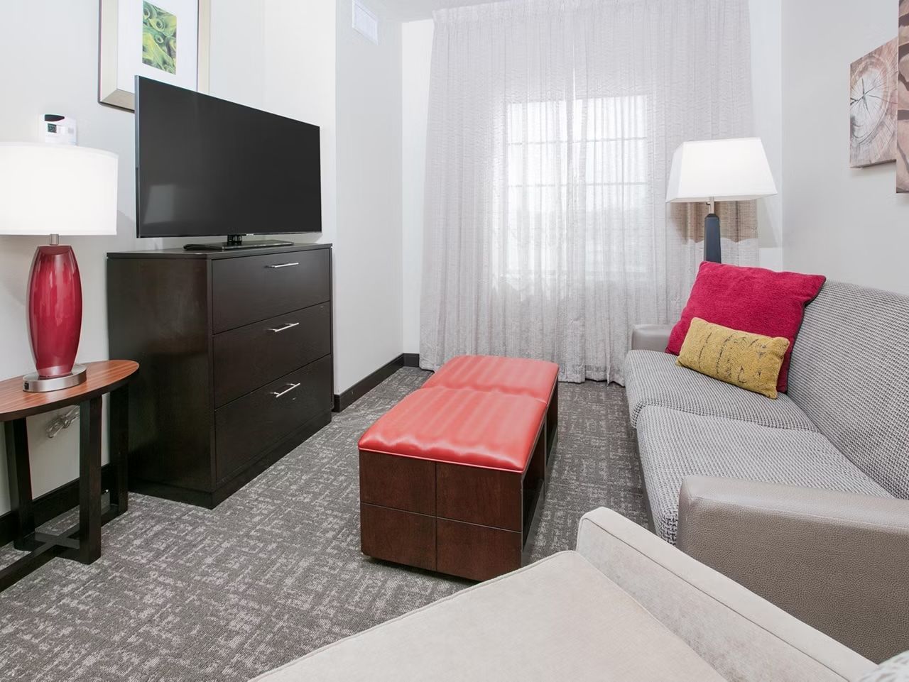 A hotel room with a flat-screen TV on a cabinet, a red ottoman, a grey sofa with a red and yellow pillow, and a white lamp.